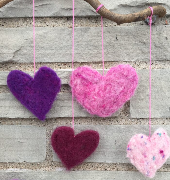 Needle Felted Hearts Kit. Four wool hearts of various colors are eacg hanging with string from a branch.
