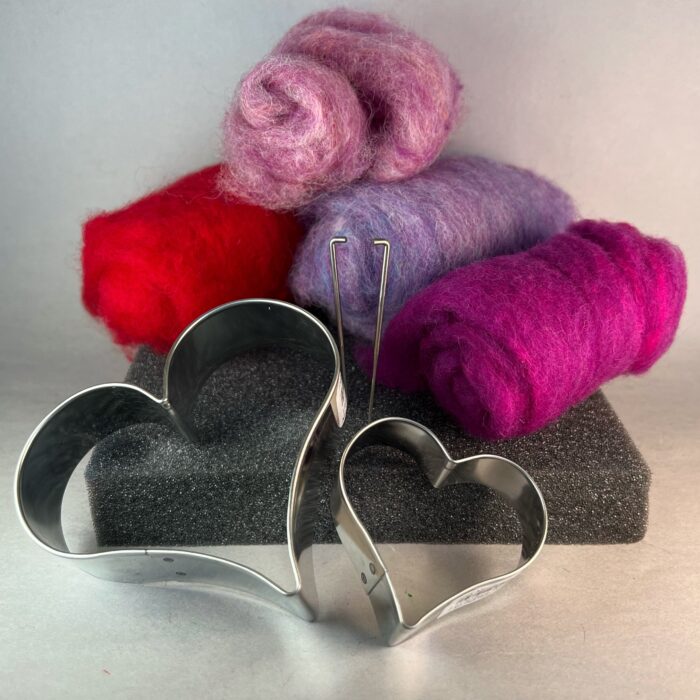 Needle Felted Hearts Kit. Four rolls of fleece in various colors are resting on a grey foam pad, with two felting needles and two heart cookie cutters.