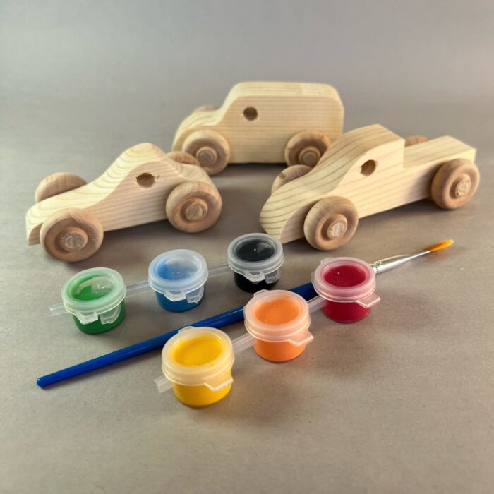 Painted Vehicles Kit. Three unpainted wood vehicles are beside six pots of paint in assorted colors and a paint brush.