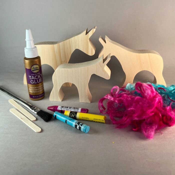 Watercolor Unicorns Kit. three wooden unicorns stand beside the contents of the craft kit: three watercolor crayons, curly mohair, glue, glue brush, craft sticks, glitter mod podge.