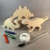 Watercolor Dinosaurs Kit. Three unpainted pine wood dinos stand beside three watercolor crayons, glitter mod podge, and a brush.