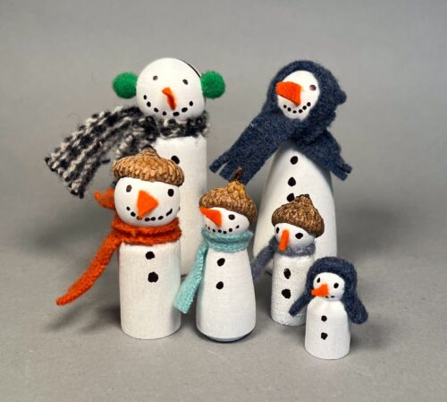 Snow Family Kit. A group of six snow people, made from wooden peg dolls painted white. Decorations include hats, scarves, ear muffs; the faces are drawn with a black marker around a felt carrot nose.