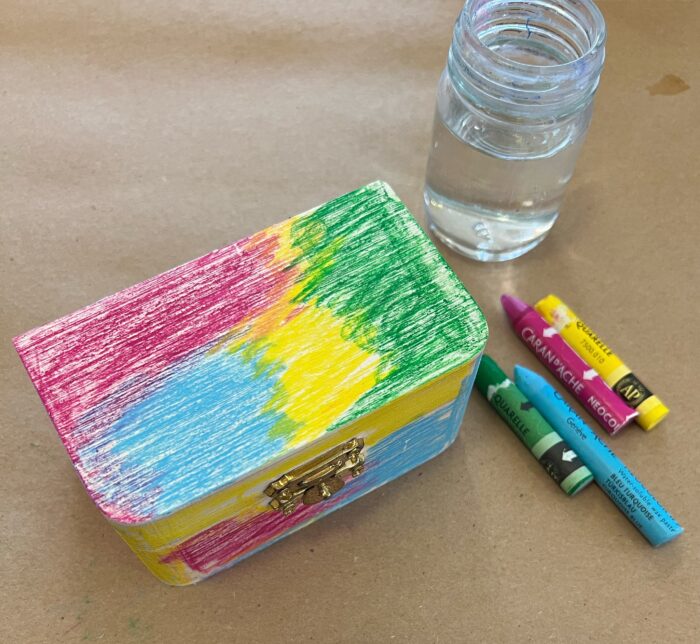Treasure Boxes Kit. Rectangular wooden box, with small jar of water and four watercolor crayons to one side. The top of the boxes has been colored with the crayons, in patches.