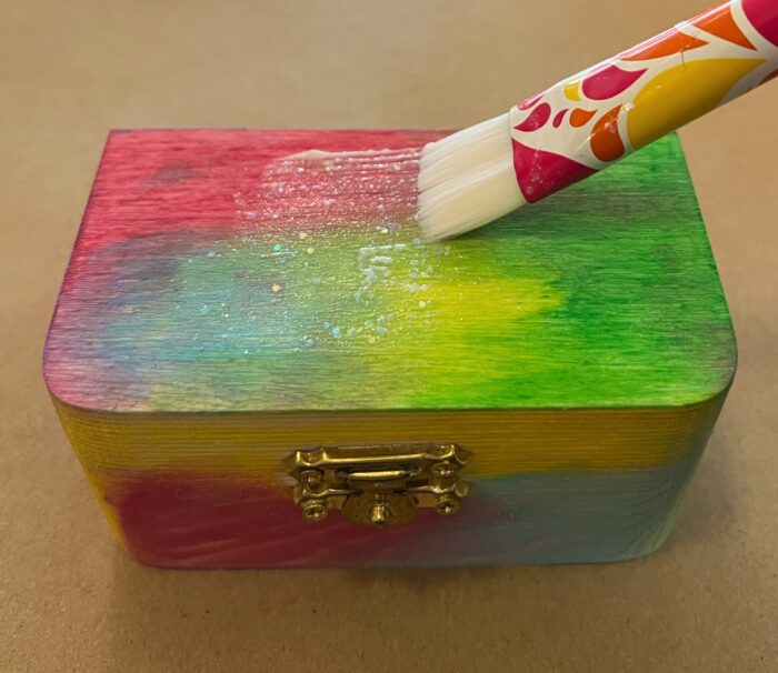 Treasure Boxes Kit. One rectangular wooden box, decorated in pink, blue, green, and yellow watercolors. A paint brush is applying a Mod Podge top coat.