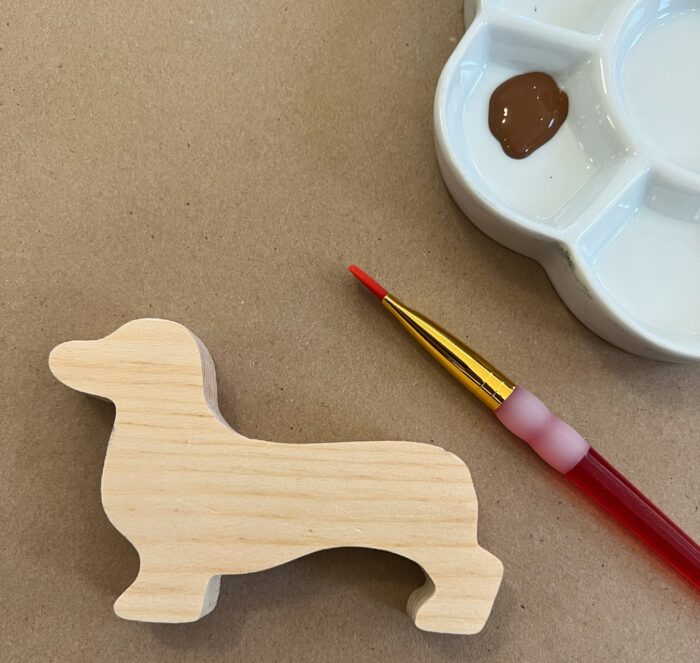 Painted Dogs Kit. Unfinished wooden dachshund cutout, with a paint brush and brown paint on the right of the dog cutout.