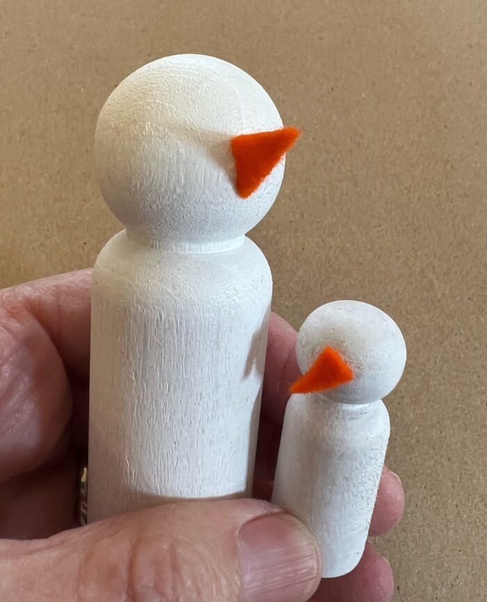 SNow Family Kit. A hand holds two wooden peg dolls, large and small. Orange felt triangles have been glued to the faces to resemble carrot noses.